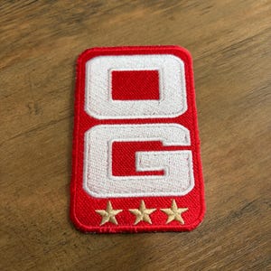 May include: A rectangular embroidered patch with a red border and background. The patch features the white letters O and G stacked vertically, with three gold-colored stars embroidered in a horizontal row along the bottom edge of the patch.