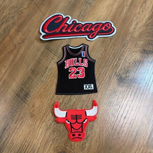 May include: Three embroidered patches featuring a black and red Chicago script logo, a black basketball jersey with the text BULLS 23 and XXL, and a red and white bull head mascot logo with black outlines, all designed for fabric application.