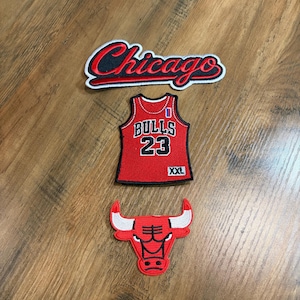 May include: Three embroidered patches featuring Chicago Bulls basketball themes. The top patch displays the word Chicago in red and black script. The middle patch is a red jersey with Bulls 23 and XXL text. The bottom patch is a red bull head logo.