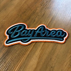 May include: Embroidered patch with the words "Bay Area" in teal script. The patch has a black background with a white and orange border, set against a wooden surface.