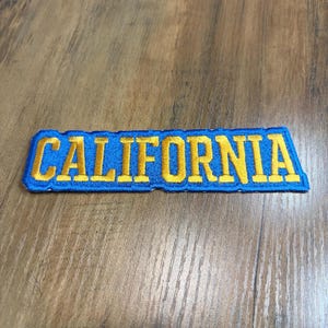 May include: A rectangular embroidered patch featuring the word CALIFORNIA in bold yellow capital letters. The text is set against a vibrant blue background with a matching blue stitched border, creating a clean and defined edge for the design.