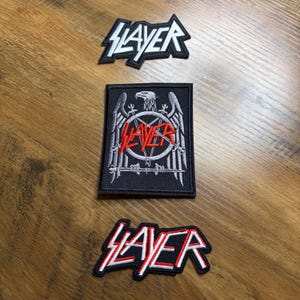 May include: Three embroidered patches with the word "SLAYER" in a bold, stylized font. One patch is a black and white logo, another features an eagle and pentagram design, and the third has a red outline.