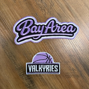 May include: Two embroidered patches on a wooden surface. The top patch reads "Bay Area" in a script font, outlined in white and black, with purple lettering. The bottom patch features a purple basketball and the word "VALKYRIES".