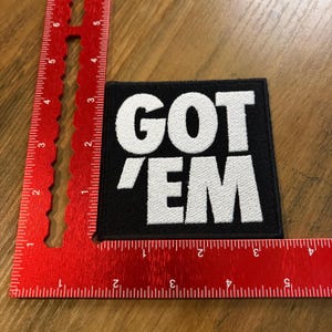 May include: Black square patch with the white embroidered text "GOT 'EM". The patch is placed on a wooden surface next to a red ruler. The patch is approximately 3 inches by 3 inches.