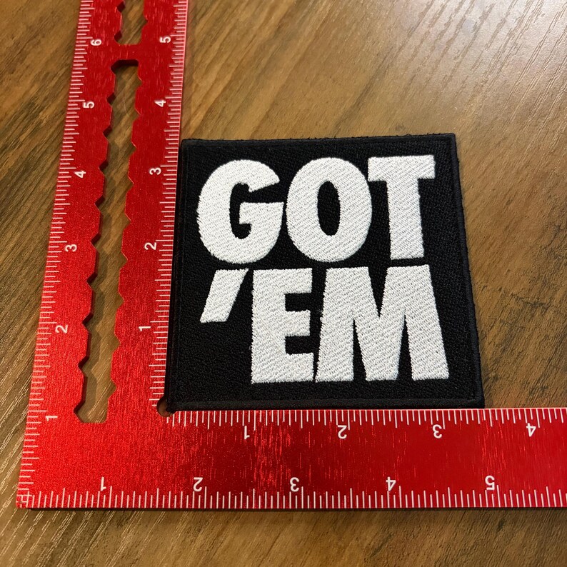 May include: Black square patch with the white embroidered text "GOT 'EM". The patch is placed on a wooden surface next to a red ruler. The patch is approximately 3 inches by 3 inches.