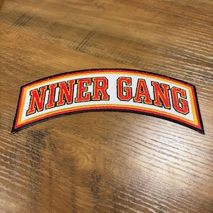 May include: A curved patch with the words "NINER GANG" in red, outlined in yellow and black, on a white background. The patch is on a wooden surface.