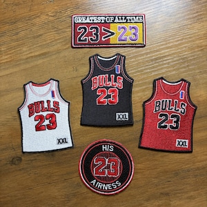 May include: A collection of embroidered patches featuring basketball-themed designs. Includes patches with the text "Bulls 23" on jerseys in white, black, and red. Also includes a patch that says "Greatest of All Time 23>23" and a round patch that says "His Airness 23".