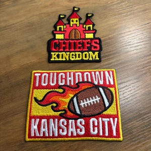 May include: Two embroidered patches. The top patch features a red and yellow castle with the text "CHIEFS KINGDOM". The bottom patch has a football with flames and the words "TOUCHDOWN KANSAS CITY".
