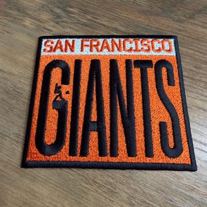May include: Embroidered patch featuring the words "SAN FRANCISCO GIANTS" in black, with an orange background. The patch has a white border and includes a small baseball player graphic. The patch is square-shaped.
