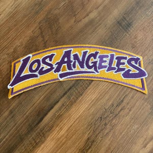 May include: Embroidered patch in a curved shape, featuring the text "Los Angeles" in purple script with a white outline, set against a yellow background. The patch is likely for apparel or accessories.