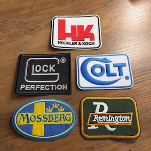 May include: Five embroidered patches with logos, including "Heckler & Koch" in red, "Glock Perfection" in white, "Colt" in blue, "Mossberg" in yellow and blue, and "Remington" in white. The patches are rectangular and oval.
