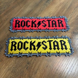 May include: Two rectangular embroidered patches featuring the text ROCK STAR in black lettering with a lightning bolt replacing the letter A. One patch has a red background and the other has a yellow background, both bordered by a grey barbed wire design.