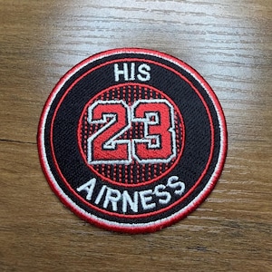 May include: Circular patch with a black background, red trim, and white and red embroidered text. The text reads "HIS 23 AIRNESS" in a bold, athletic font. The patch is likely for clothing or accessories.