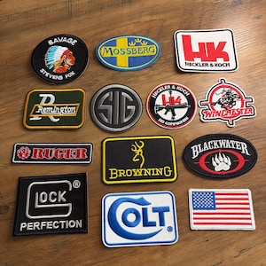 May include: A collection of embroidered patches with various logos and text. Patches include "SAVAGE", "MOSSBERG", "Remington", "RUGER", "BROWNING", "BLACKWATER", "COLT", "LOCK PERFECTION", "HECKLER & KOCH", and a US flag.