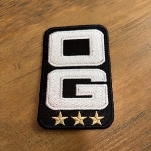 May include: A rectangular embroidered patch with a black background and white stitched letters spelling OG. Below the letters are three small, gold-colored embroidered stars arranged in a horizontal row. The patch has a black stitched border.