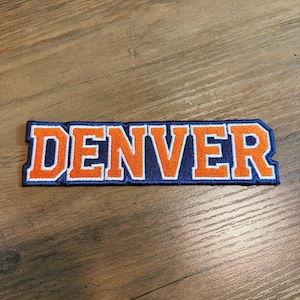 May include: A rectangular embroidered patch featuring the word DENVER in bold orange block letters with a white outline, set against a dark navy blue background. The lettering has a textured, stitched appearance with a distinct border.