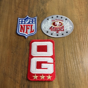 May include: Three embroidered patches: a shield-shaped NFL logo in red, white, and blue; an oval San Francisco 49ers patch with a city skyline, red and gold logo, and black stars; and a rectangular red patch with white letters OG and three gold stars.