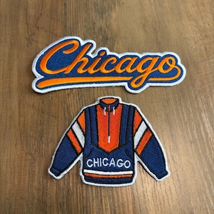 May include: Two embroidered patches featuring a Chicago theme. The top patch displays the word Chicago in orange script with a blue outline. The bottom patch is a blue, orange, and white pullover jacket with the word Chicago printed in white.