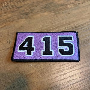 May include: A rectangular embroidered patch featuring the black numbers 415 centered on a light purple background. The patch has a thick black border surrounding the purple field, with white outlines separating the black numbers from the purple fabric.