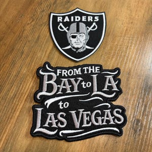 May include: Two embroidered patches. The top patch is a black and white shield-shaped Raiders logo featuring a football helmet and crossed swords. The bottom patch is black with white embroidered text reading FROM THE BAY to LA to LAS VEGAS.