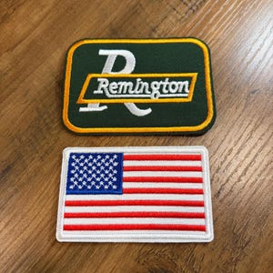 May include: Two embroidered patches. One is a green rectangle with the word "Remington" in white and gold. The other is a rectangular patch of the American flag with red and white stripes and a blue field of stars.