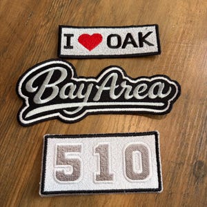 May include: Three embroidered patches. The top rectangular patch is white with black text reading I, a red heart symbol, and OAK. The middle patch is black with white cursive text reading Bay Area. The bottom rectangular patch is white with the number 510.