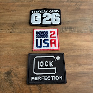 May include: Three rectangular embroidered patches. The top patch is black with white text reading EVERYDAY CARRY G26. The middle patch is white with a blue American flag graphic, red text 2, and blue text USA. The bottom patch is black with white text GLOCK PERFECTION.