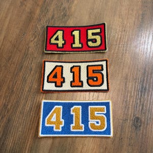 May include: Three rectangular embroidered patches, each featuring the number 415. The top patch is red with gold numbers, the middle is cream with orange numbers, and the bottom is blue with gold numbers, all outlined in black thread.