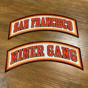 May include: Two arched patches with the words "SAN FRANCISCO" and "NINER GANG" in red, outlined in white, yellow, and black. The patches are likely for clothing or accessories.