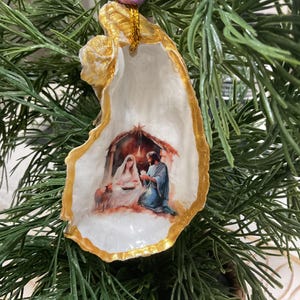 May include: A decorative oyster shell ornament featuring a painted nativity scene with Mary, Joseph, and the infant Jesus. The shell edges are painted gold, and it hangs from a gold string topped with a single purple bead.