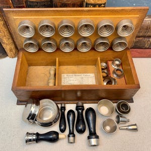 May include: An antique wooden box with a variety of medical tools. The box contains metal tools, including circular cutters, funnels, and stamps. The box is open, revealing the tools inside. Some tools have wooden handles. The box is sitting on a light-colored surface.
