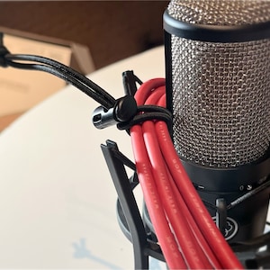 May include: A black studio microphone with a silver mesh head. Red cables are bundled together with a black cord and clip. The microphone is mounted on a black stand, with a white surface in the background.