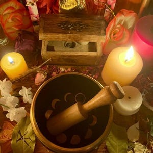 Pode incluir: Uma composi&ccedil;&atilde;o espiritual com uma ta&ccedil;a cantora preta com desenhos de fases lunares, um malho de madeira e v&aacute;rios cristais. Velas acesas, uma pequena caixa de madeira e uma bolsa dourada criam um ambiente m&iacute;stico.