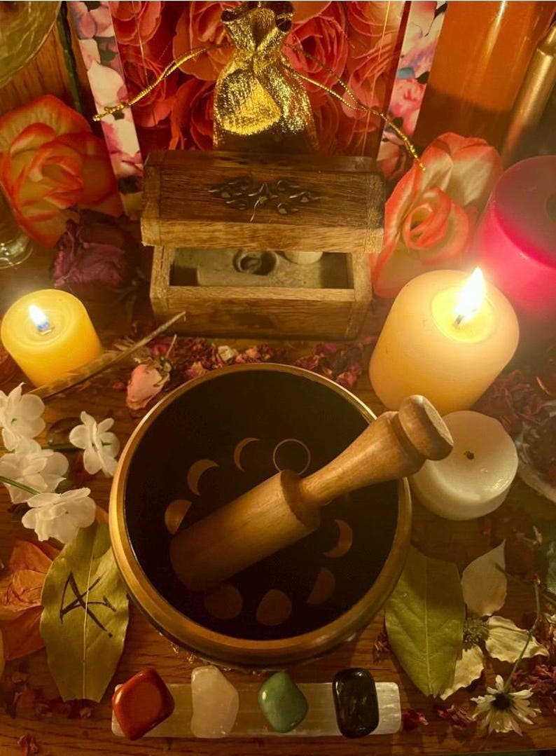 Pode incluir: Uma composi&ccedil;&atilde;o espiritual com uma ta&ccedil;a cantora preta com desenhos de fases lunares, um malho de madeira e v&aacute;rios cristais. Velas acesas, uma pequena caixa de madeira e uma bolsa dourada criam um ambiente m&iacute;stico.
