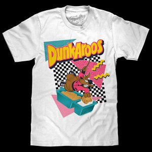 May include: A white short-sleeve t-shirt featuring a graphic print of the Dunkaroos logo in yellow and red. The design includes a cartoon kangaroo wearing a pink jacket, a teal snack container, and geometric patterns in black, white, and pink.