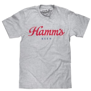May include: A heather gray short-sleeve t-shirt featuring the red cursive text Hamm's and the blue block text BEER printed on the chest. The inner neck label displays the Tee Luv logo. The shirt has a crew neckline and a standard fit.