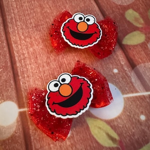 May include: Two red glitter hair bows with Elmo character embellishments. Each bow features a red bow shape with a glittery texture and a smiling Elmo face in the center. The bows are set against a reddish-brown background.