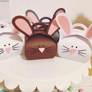 May include: Several bunny-shaped gift boxes in white and brown, with pink inner ears and pink noses. The boxes have black whiskers and handles. They are arranged on a white, scalloped-edge cake stand, suggesting an Easter theme.