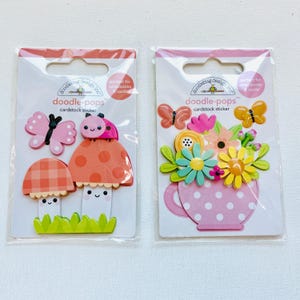 May include: Two packages of Doodlebug Design cardstock stickers. One features a mushroom and butterfly design, the other a floral arrangement in a pink polka-dot teacup. The packages are sealed in clear plastic.