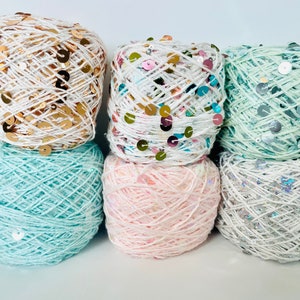May include: Six balls of yarn in various colors, each adorned with sequins. Colors include white with gold sequins, white with multicolored sequins, mint green with silver sequins, light blue, pink, and white with iridescent sequins. Ideal for crafting.