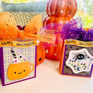 May include: Two rectangular Halloween treat boxes with orange and purple tulle bows. The left box features a pumpkin mug graphic with a straw and the text HAPPY HALLOWEEN. The right box has a spider web window filled with colorful beads and a spider.