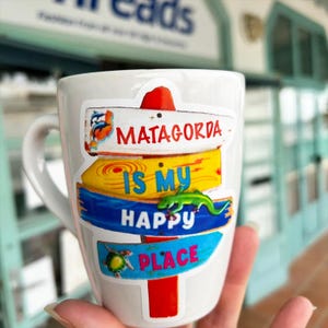 May include: White ceramic mug with a handle, featuring a colorful design of Matagorda signs. The signs read "Matagorda Is My Happy Place" with a green lizard and a turtle. The mug is held in a hand.