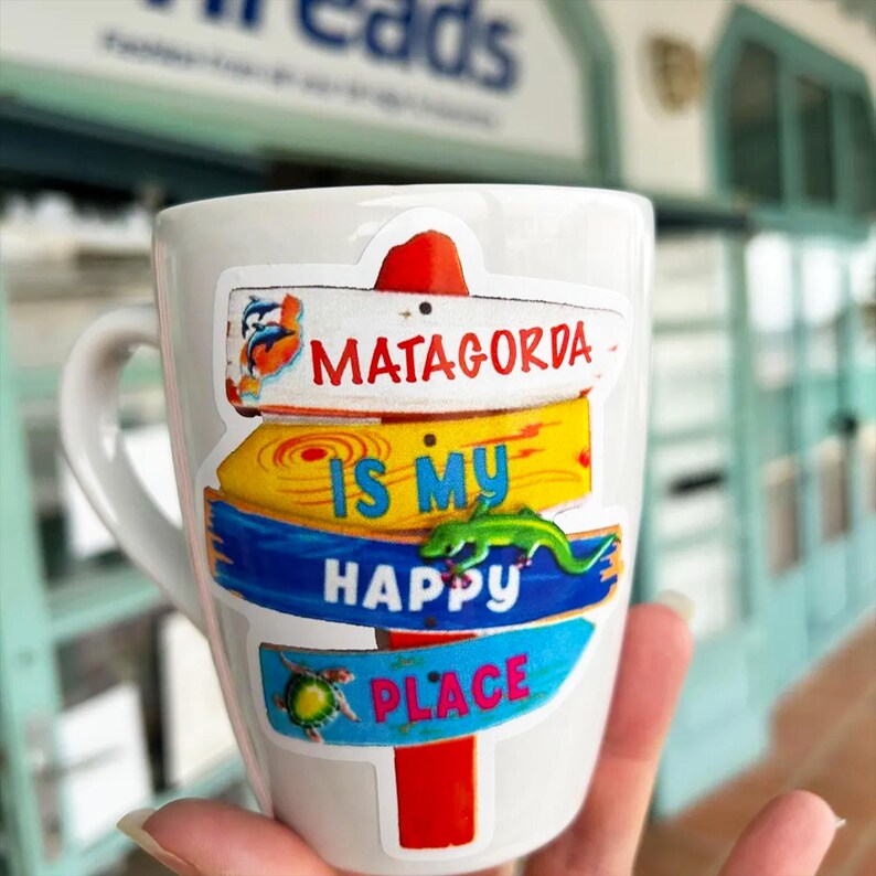 May include: White ceramic mug with a handle, featuring a colorful design of Matagorda signs. The signs read "Matagorda Is My Happy Place" with a green lizard and a turtle. The mug is held in a hand.