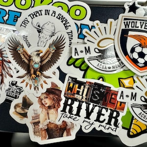 May include: A collection of assorted vinyl stickers featuring various designs including a skull with a feathered headdress, a bald eagle, a woman in a corset, a soccer team crest labeled Wolverines, and text graphics reading Whiskey River and A-M Acha Morfaw.