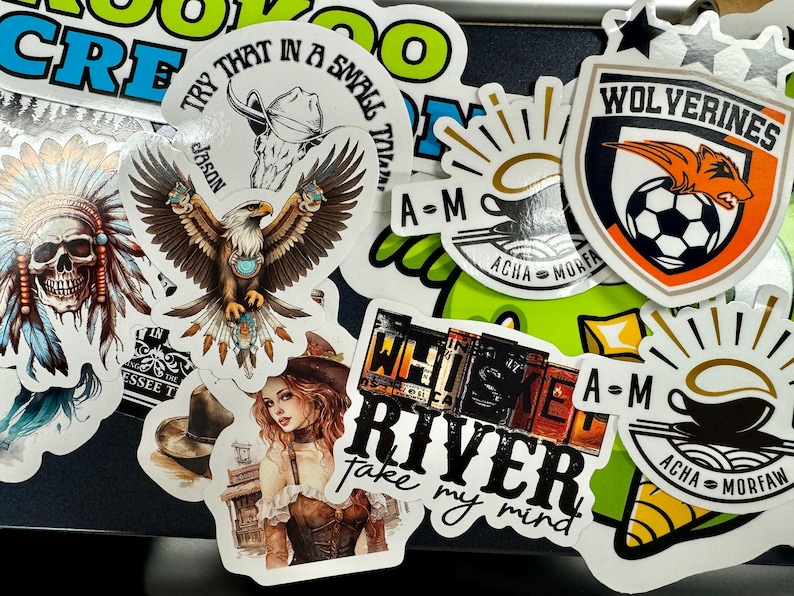 May include: A collection of assorted vinyl stickers featuring various designs including a skull with a feathered headdress, a bald eagle, a woman in a corset, a soccer team crest labeled Wolverines, and text graphics reading Whiskey River and A-M Acha Morfaw.