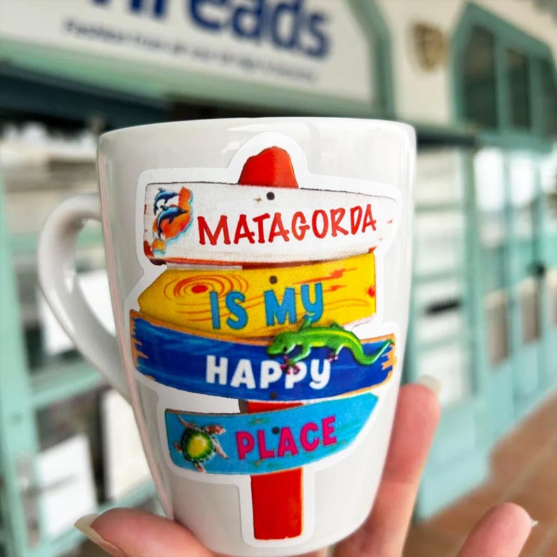 May include: A white ceramic mug featuring a colorful sticker of a multi-tiered signpost. The sign reads MATAGORDA IS MY HAPPY PLACE in red, blue, and yellow text, decorated with illustrations of two dolphins, a green lizard, and a sea turtle.