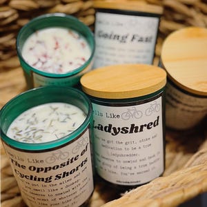 May include: Three glass candles in dark green containers with light-colored wooden lids. The labels feature bicycle icons and text including Smells Like Ladyshred and The Opposite of Cycling Shorts. The wax is white with visible botanical inclusions.