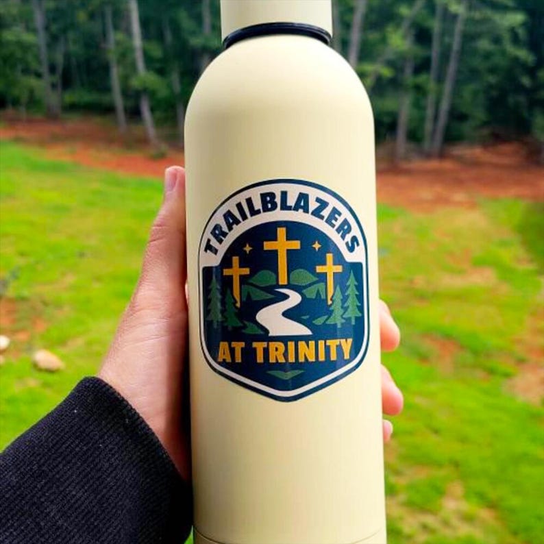 May include: A cream-colored metal water bottle featuring a graphic sticker with the text TRAILBLAZERS AT TRINITY. The design includes three yellow crosses, a winding white path, green pine trees, and a dark blue background shaped like a shield.