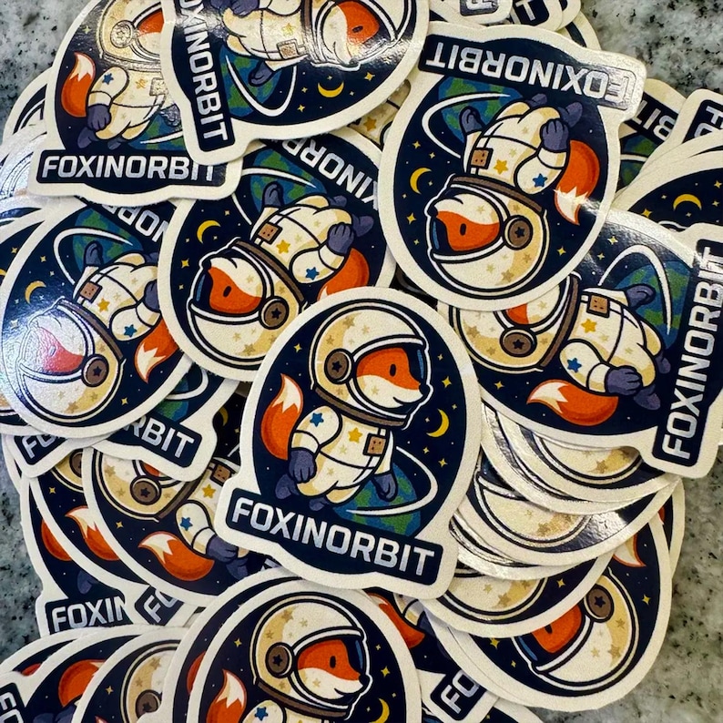 May include: A circular sticker featuring a cartoon fox wearing a white astronaut suit with colorful star accents. The fox is floating in space against a dark blue background with a crescent moon and a partial view of Earth. The text FOXINORBIT is printed.