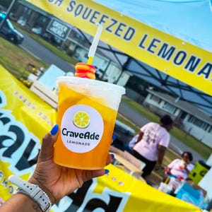 May include: A clear plastic cup filled with bright orange lemonade, featuring a white circular label with a yellow lemon graphic and the text CraveAde LEMONADE. A white straw with a red and yellow gummy candy ring sits in the drink.